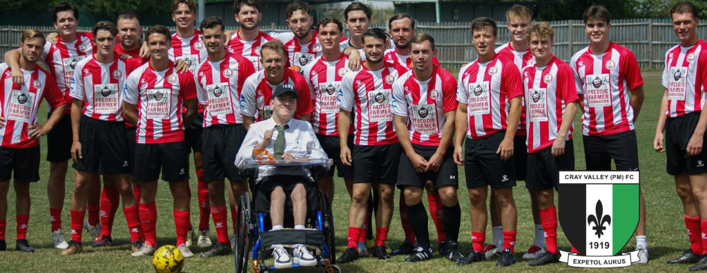 Group photo on a football pitch showing members of Team Freddie Farmer Foundation wearing red-and-white striped shirts, black shorts and red socks. A wheelchair user is positioned at the front centre of the group. A football rests on the grass in the foreground, with trees and stadium fencing visible in the background. The Cray Valley (PM) FC club crest appears in the lower-right corner of the image.
